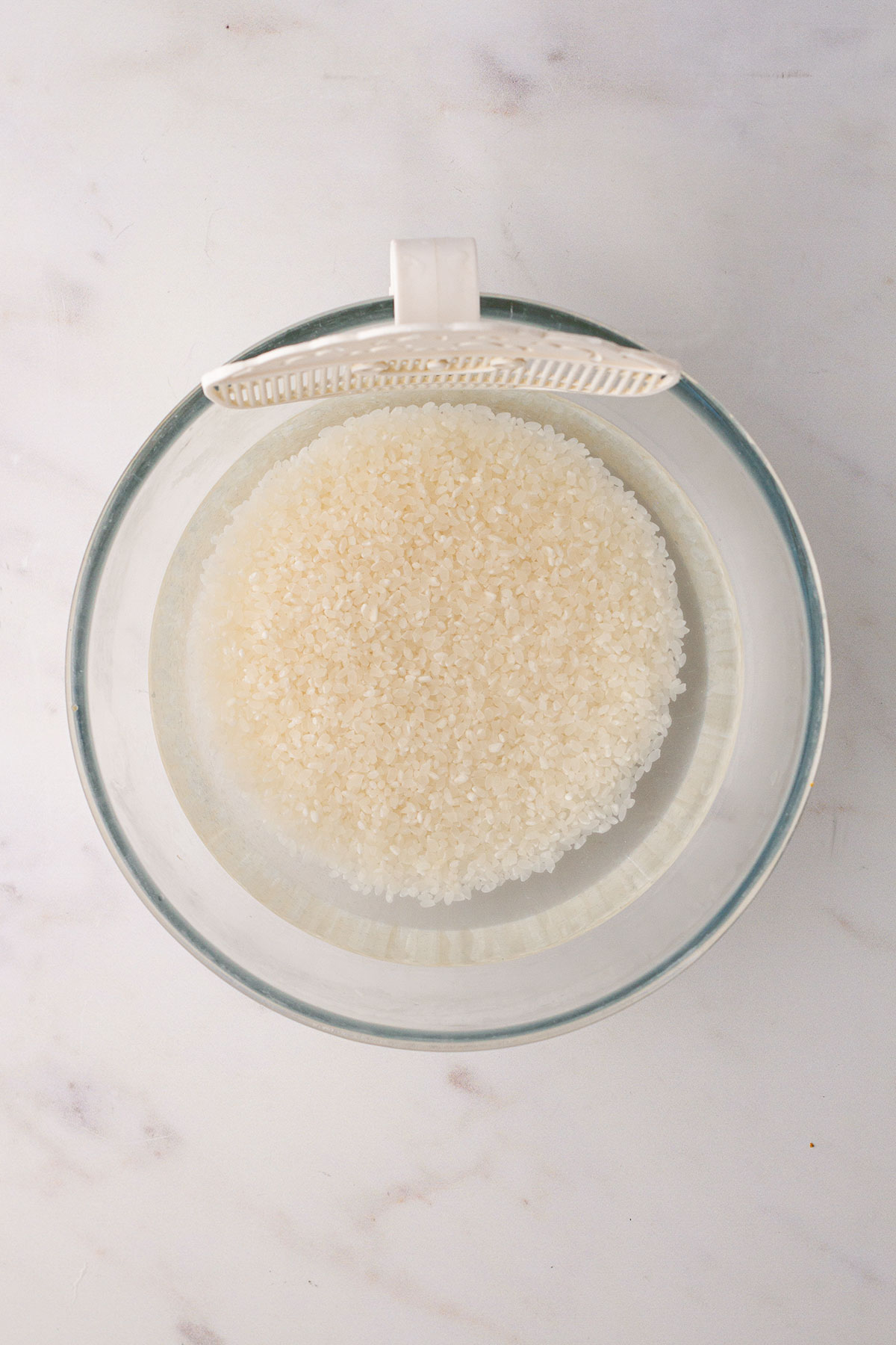 Washing the rice in a glass bowl with a rice strainer clip attached, on a white background.