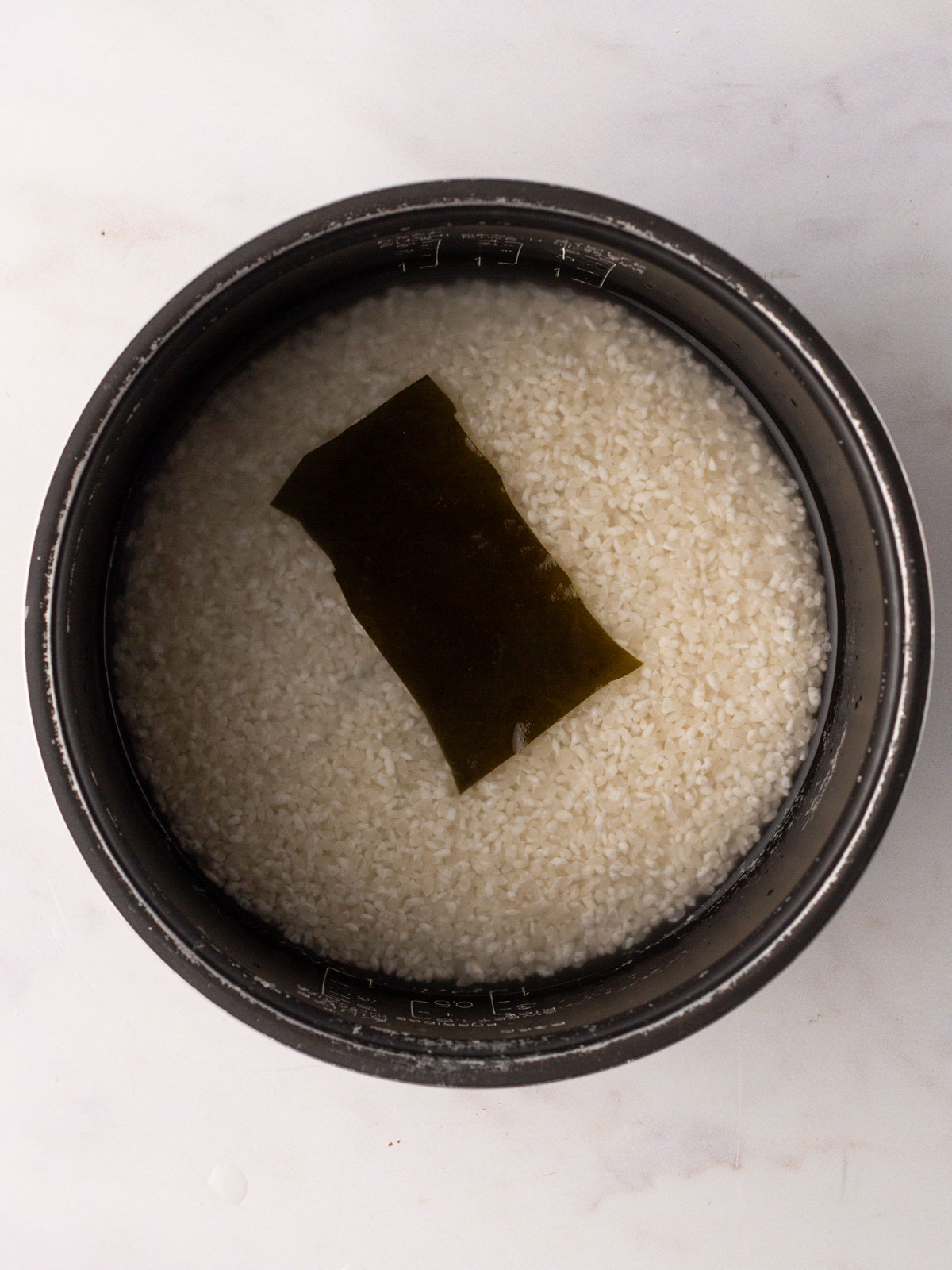 A rice cooker pan on a white background containing the rinsed rice, water and kombu.