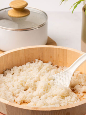 The cooked sushi rice in a light brown ohitsu container with a white spoon.