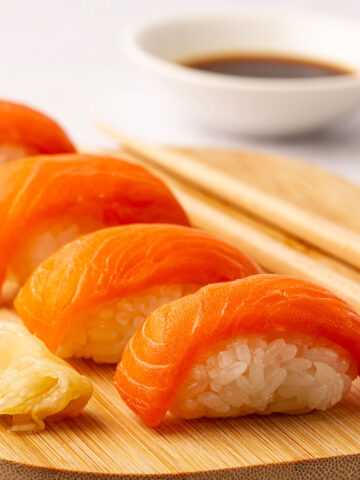 Four pieces of salmon nigiri on a wooden board with chopsticks and a white bowl containing soy sauce in the background.