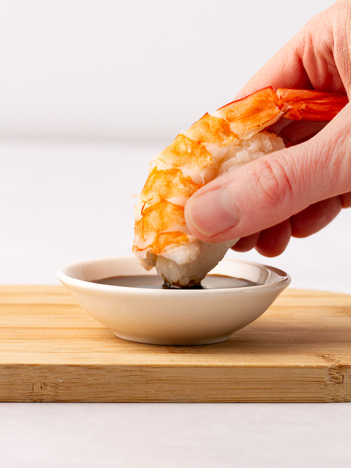 A shrimp nigiri being dipped into a white bowl of soy sauce on a wooden board.