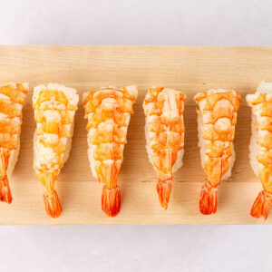 Six finished shrimp nigiri on a wooden board on a white background.