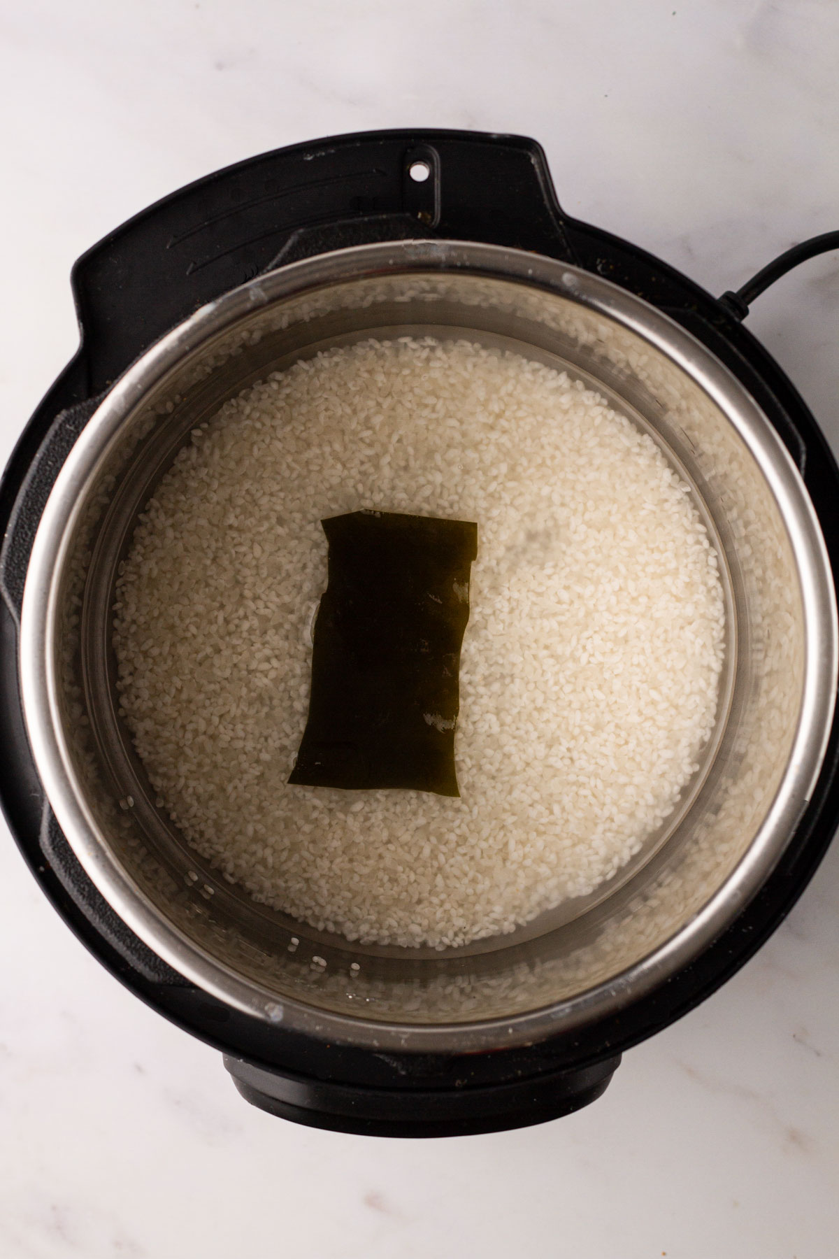 An Instant Pot on a white background containing the rinsed rice, water and kombu.