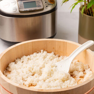 The cooked sushi rice in a light brown ohitsu container with a white spoon on a bamboo placement mat with the rice cooker and a potted plant behind.