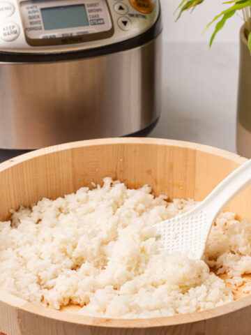 The cooked sushi rice in a light brown ohitsu container with a white spoon on a bamboo placement mat with the rice cooker and a potted plant behind.