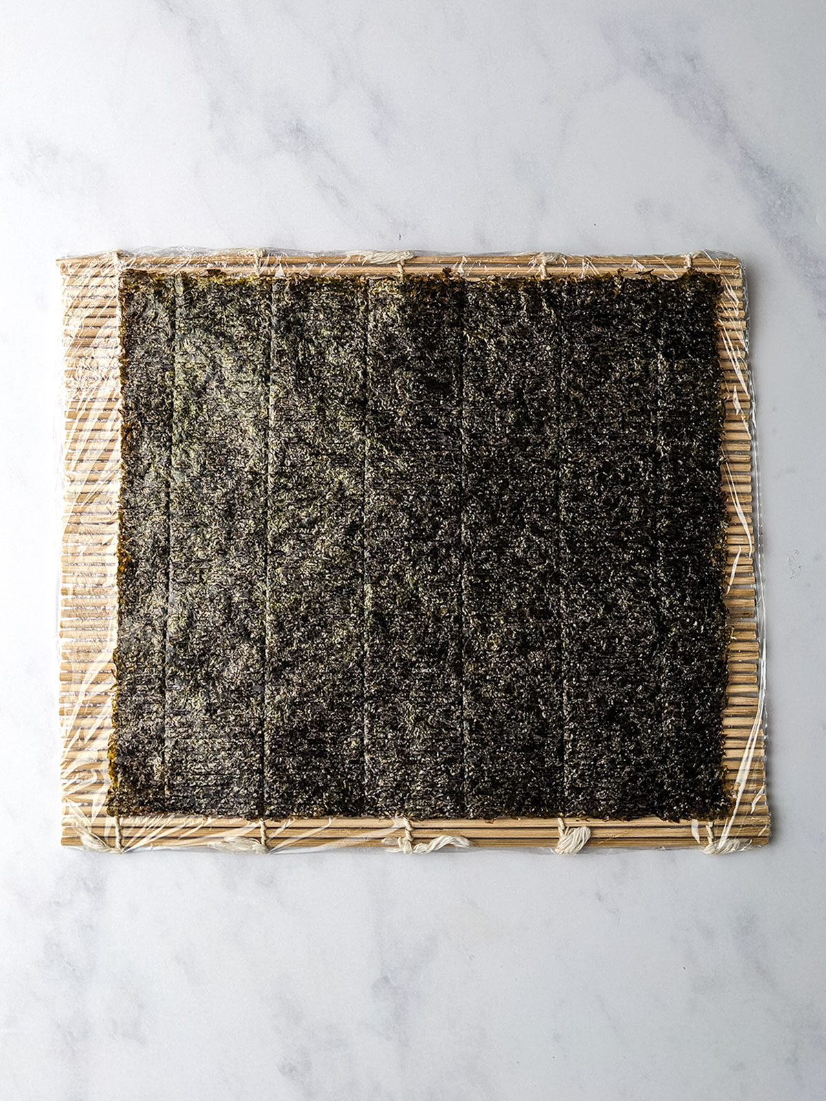 The nori sheet placed rough side facing up on top of the bamboo mat wrapped in plastic wrap.