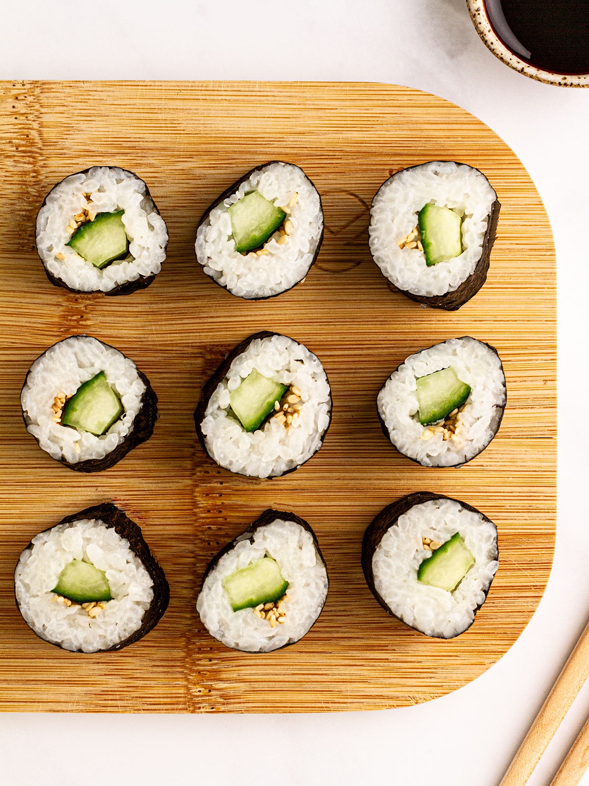 Nine completed kappa maki cucumber rolls on a wooden board on a white background with chopsticks and a bowl of soy sauce.