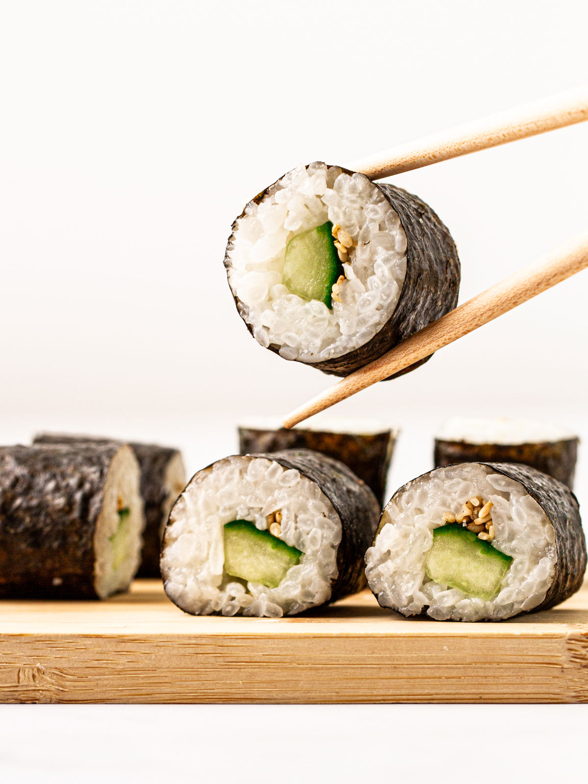 Chopsticks holding a kappa maki cucumber roll above more rolls on a wooden board on a white background.