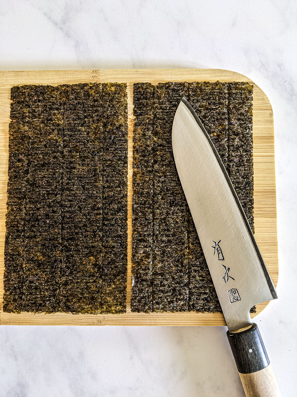 Two halves of nori sheet cut to fit the size of the rolls showing a knife on a bamboo rolling mat on a wooden board on a white background.