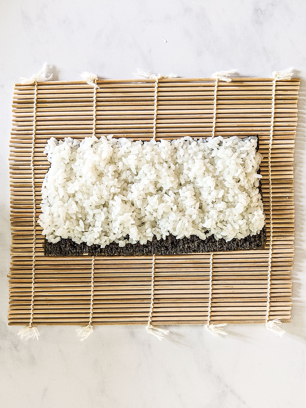 A layer of sushi rice spread over the nori on a bamboo rolling mat on a white background.