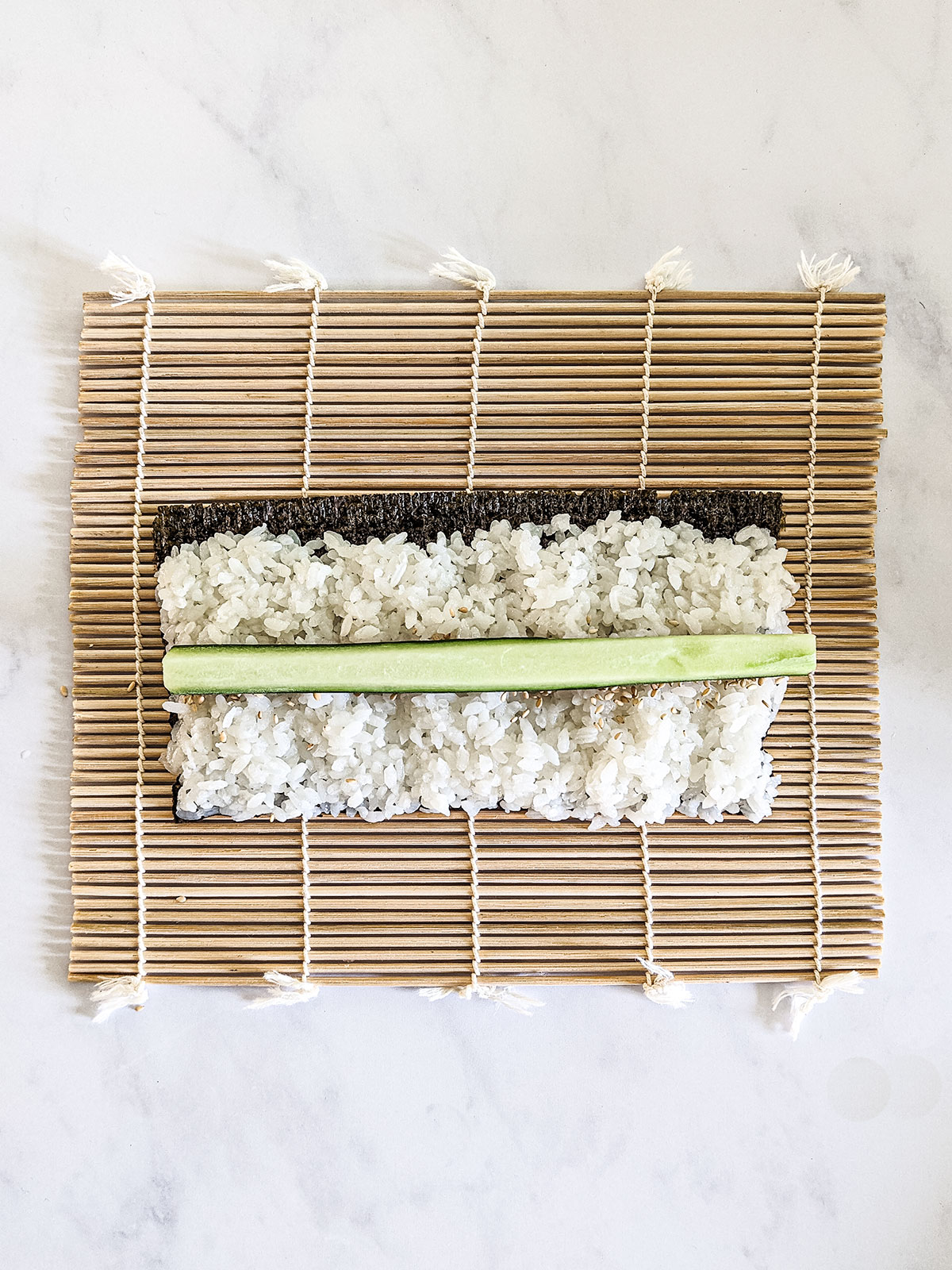 Toasted sesame seeds sprinkled across the middle of the rice layer on the nori with a stick of cucumber on top, on a bamboo rolling mat on a white background.