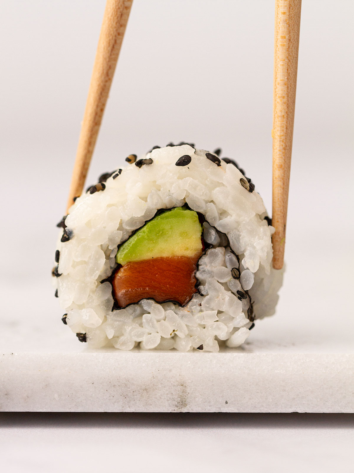 A closeup of a salmon and avocado roll with chopsticks.